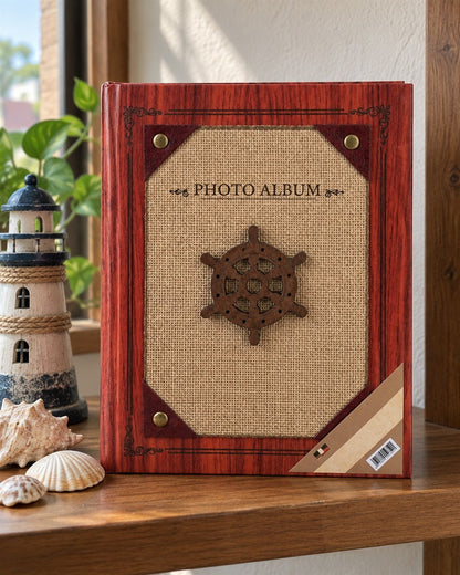 Álbum de fotos de tapa dura para 200 fotos con temática náutica: portada con acabado imitación madera caoba rojiza y marco decorativo, panel central de tela tipo yute color arena con un timón de barco de madera oscura en su lugar de uso