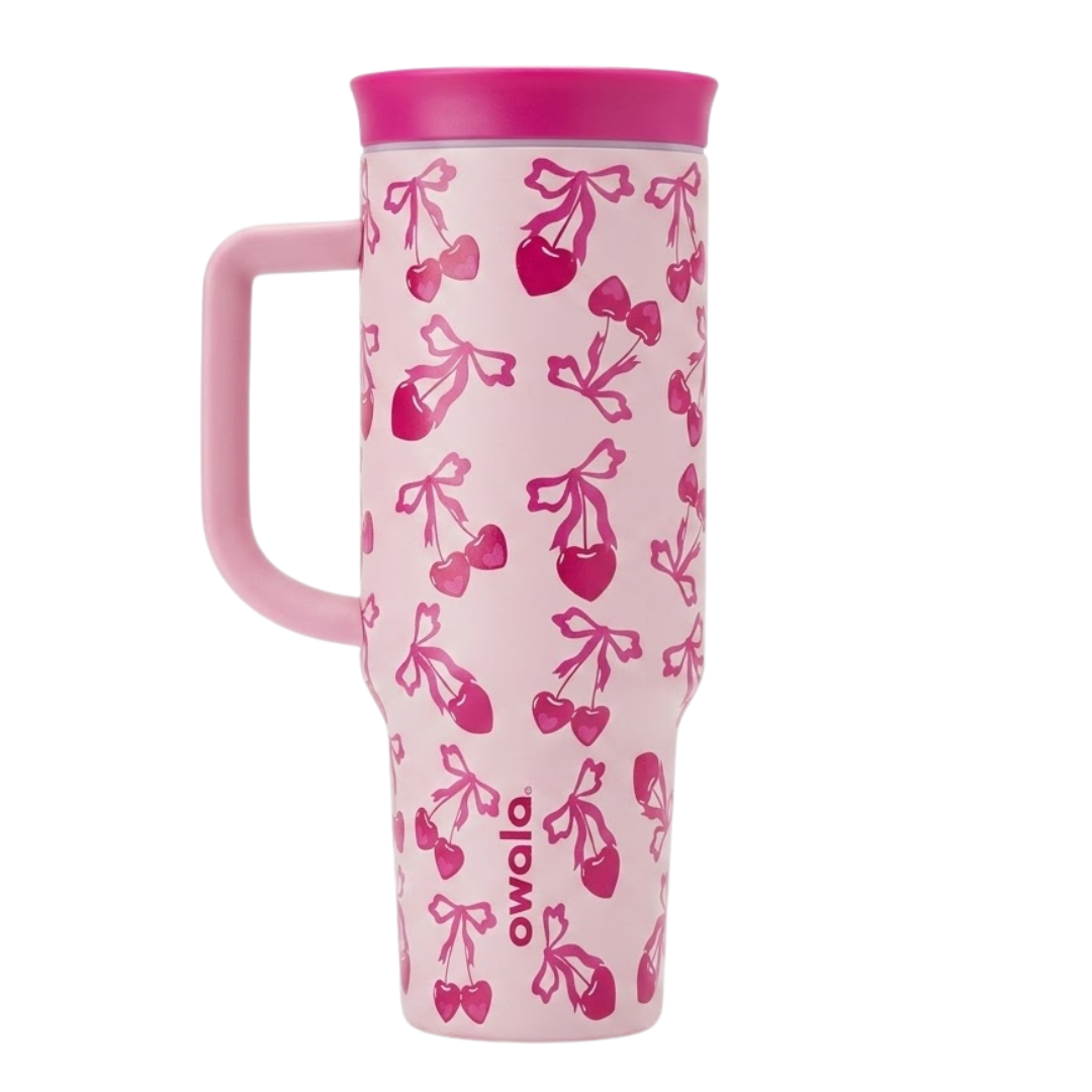 Vaso térmico Owala de 40 onzas en color rosa pastel con un patrón de cerezas en forma de corazón y lazos en color fucsia. El diseño incluye una tapa fucsia vibrante, asa ergonómica rosa y base cónica. Logotipo de Owala en vertical en la parte inferior en color fucsia.