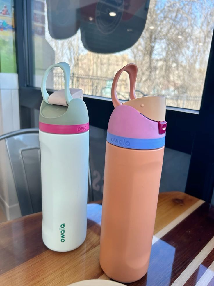 Dos botellas de agua Owala FreeSip de 24 onzas de acero inoxidable. La botella de la izquierda (variante Neo Sage) es de color celeste pastel muy claro con el logotipo de Owala en verde oscuro y una colorida tapa en tonos verde oliva, fucsia y menta. La botella de la derecha (variante Coastal Mist) es de color durazno o salmón mate con el logotipo en café oscuro y una tapa en tonos morado, rosa y azul cielo.