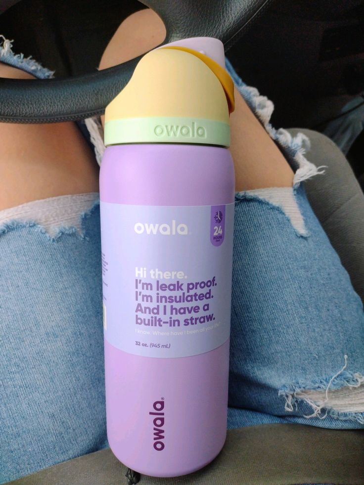 Botella de agua Owala FreeSip de 32 onzas El cuerpo es de color lavanda o lila mate con una banda protectora original que detalla sus funciones en letras moradas. La tapa presenta una combinación estética de tonos pastel: la base de la rosca es verde menta claro con la marca Owala en relieve, la cubierta principal es de color crema o amarillo suave, y el botón de apertura junto al asa de transporte son de un tono naranja apagado o mostaza.
