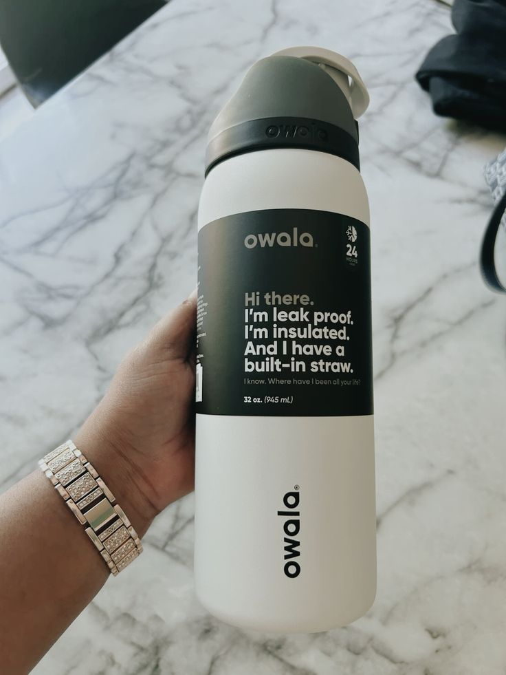 Botella de agua de acero inoxidable Owala FreeSip de 32 onzas en color blanco. El termo tiene su etiqueta original negra que detalla sus funciones principales. La tapa presenta un diseño monocromático con una base negra donde resalta la marca Owala, una cubierta superior gris oscuro y un asa de transporte en gris claro o blanco. El logotipo de Owala está impreso verticalmente en negro cerca de la base.