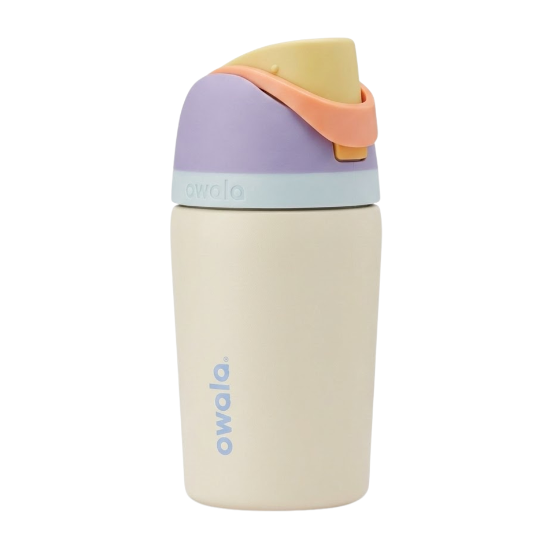 Botella térmica de 12 onzas para niños modelo Owala Kids FreeSip en color crema o beige suave. La tapa combina tonos lila, un asa en color melocotón claro y una banda en color menta muy suave. Logotipo de Owala en color lila vertical sobre el cuerpo crema.