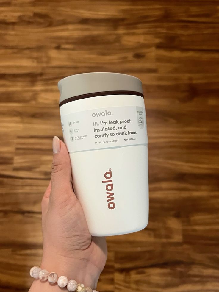 Vaso térmico compacto para café Owala SmoothSip de 12 onzas siendo sostenido por una mano frente a un fondo de suelo de madera. El cuerpo es de color crema suave mate con la marca Owala impresa verticalmente en un tono café terracota. Tiene su etiqueta original blanca que detalla sus funciones. La tapa es de color gris claro con una línea intermedia en café oscuro.