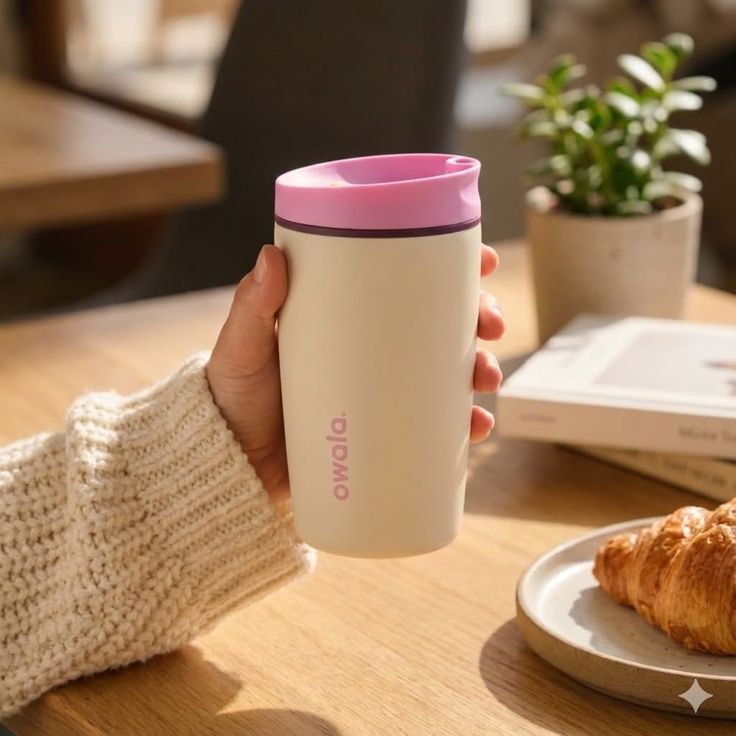 Vaso térmico para café Owala SmoothSip de 20 onzas siendo sostenido por una mano. El cuerpo es de color crema suave o arena mate, con el logotipo de Owala impreso verticalmente en color rosa. La tapa es de color rosado pastel con una sutil banda intermedia en color uva oscuro y cuenta con una boquilla diseñada para un flujo suave de bebidas calientes.