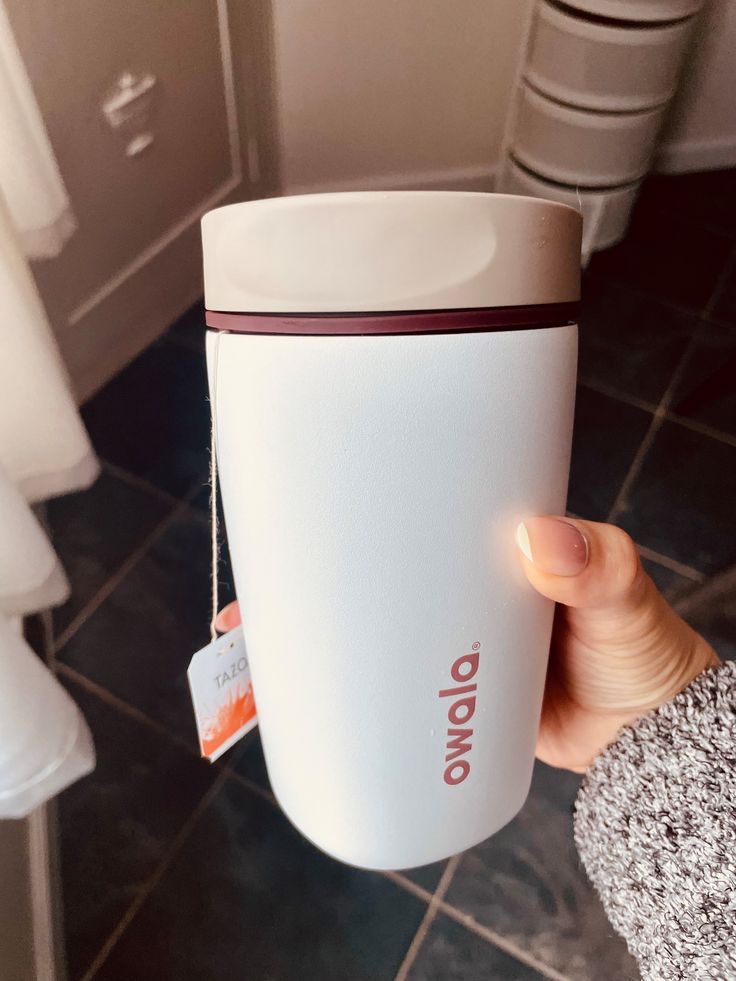 Vaso térmico mediano para café o té Owala SmoothSip de 20 onzas. El cuerpo de acero inoxidable es de color blanco con el logotipo de Owala impreso de forma vertical en un tono café terracota. De la tapa de color gris claro sobresale la cuerda con la etiqueta de una bolsa de té Tazo.