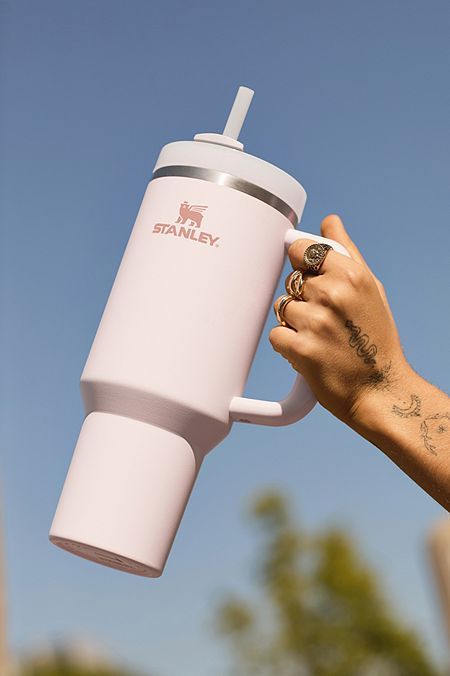 Termo grande Stanley Quencher H2.0 de 40 onzas en color rosa pastel muy claro (Rose Quartz) siendo sostenido en el aire frente a un cielo azul despejado. El termo muestra el logotipo de Stanley en un elegante tono oro rosa o café suave.