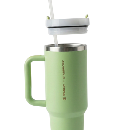 Vaso térmico Stanley de 40 onzas en color verde, perteneciente a una colaboración especial con Starbucks. En el lateral, presenta un grabado vertical elegante que dice "STANLEY + STARBUCKS" junto al icono del oso alado de Stanley, todo en un tono dorado suave o bronce. El asa y el cuerpo son del mismo tono verde lima, con una tapa transparente que tiene un cierre superior y pajilla en verde a juego.