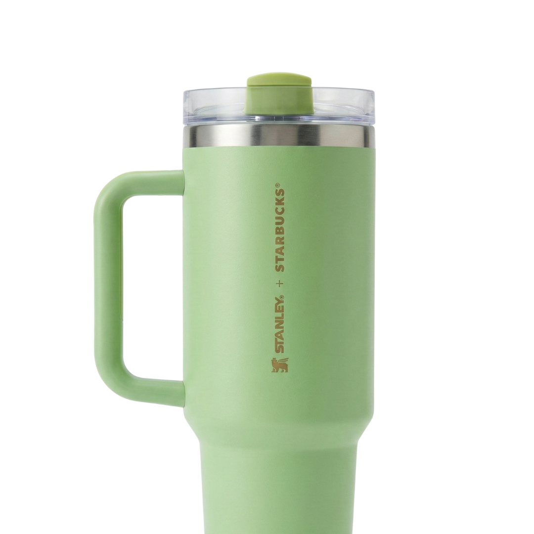Vaso térmico Stanley de 40 onzas en color verde, perteneciente a una colaboración especial con Starbucks. En el lateral, presenta un grabado vertical elegante que dice "STANLEY + STARBUCKS" junto al icono del oso alado de Stanley, todo en un tono dorado suave o bronce. El asa y el cuerpo son del mismo tono verde lima, con una tapa transparente que tiene un cierre superior y pajilla en verde a juego.
