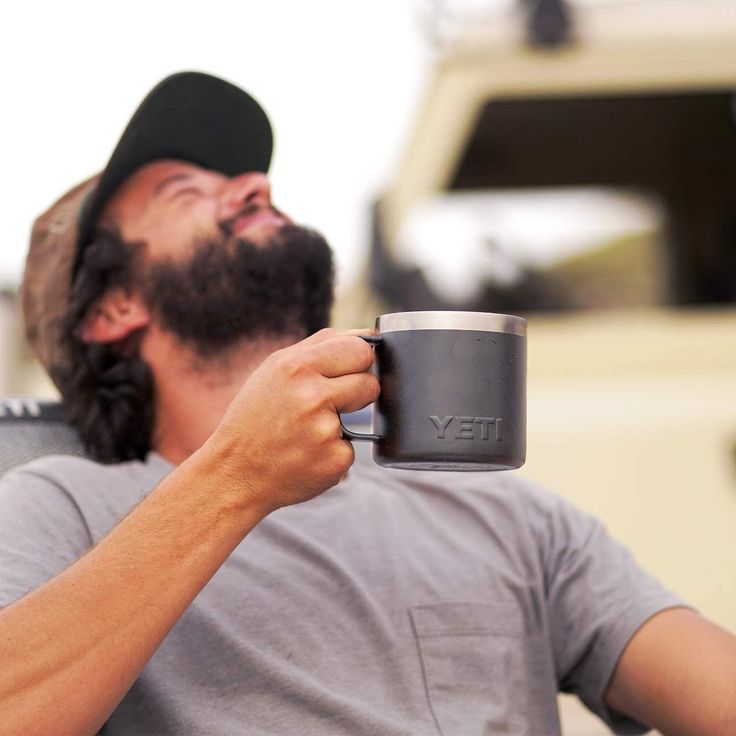 Taza térmica tipo campamento Yeti Rambler en color negro mate con asa integrada. El cuerpo tiene un acabado de pintura en polvo negra de alta resistencia con el logotipo de YETI grabado en relieve en la parte frontal. Cuenta con un borde superior de acero inoxidable expuesto.