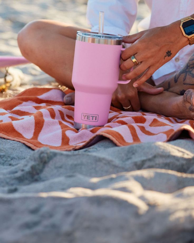 aso térmico con asa Yeti Rambler Travel Mug de 30 onzas en color rosado brillante (Power Pink) con acabado mate. Está colocado sobre una toalla playera en la arena. El frente muestra el logotipo clásico de YETI en un recuadro de acero inoxidable cerca de la base. Cuenta con una tapa transparente con pajilla y un asa integrada del mismo tono rosado para un agarre firme.